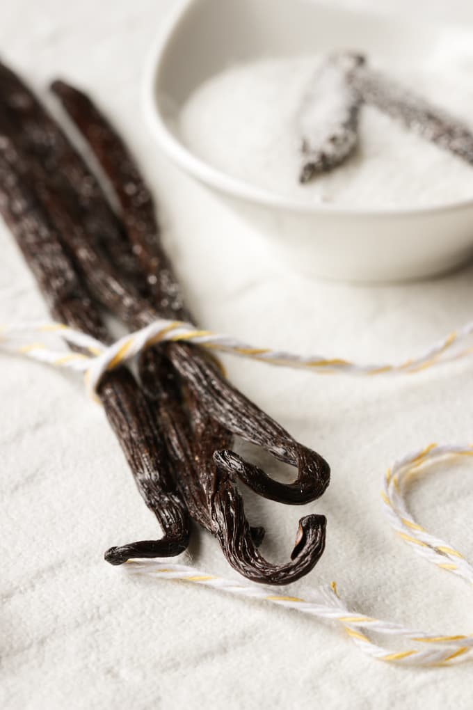 Five vanilla beans tied with a yellow and white ribbon and a white bowl in the background.