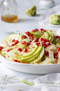 Green apple and fennel salad topped with pomegranate arils on a white plate.