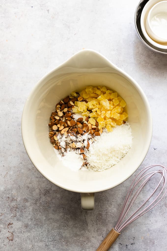 A white mixing bowl with flour, almonds, coconut and pineapple pieces.