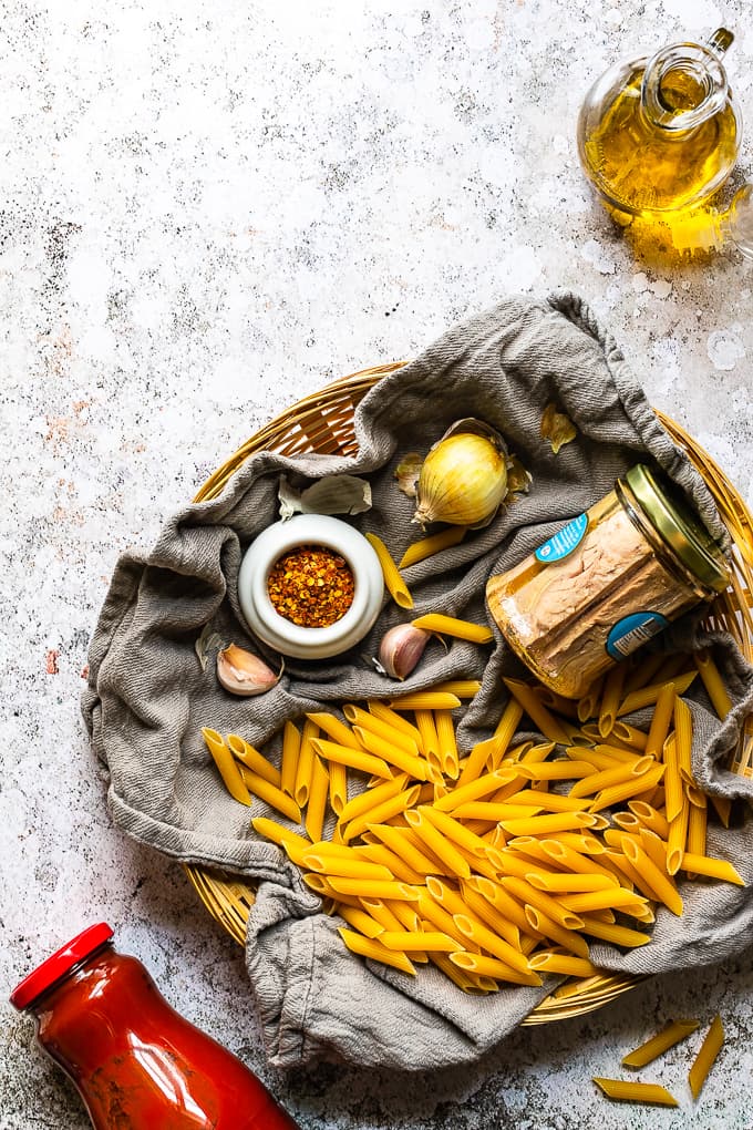 A wicker basket filled dry penne pasta, a jar of tuna fillet, onion, garlic and spices.