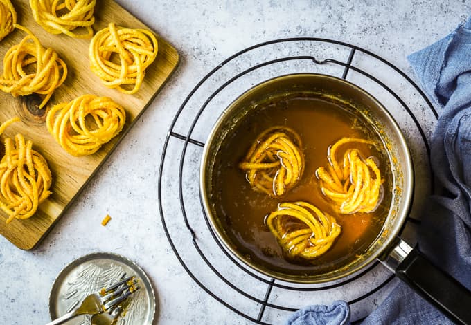 Three deep fried twirled pasta in a pot of warmed honey on a black rack.