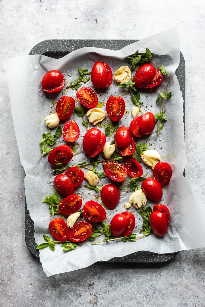 Cherry tomatoes, garlic and oregano tossed with olive oil on a grey baking tray.