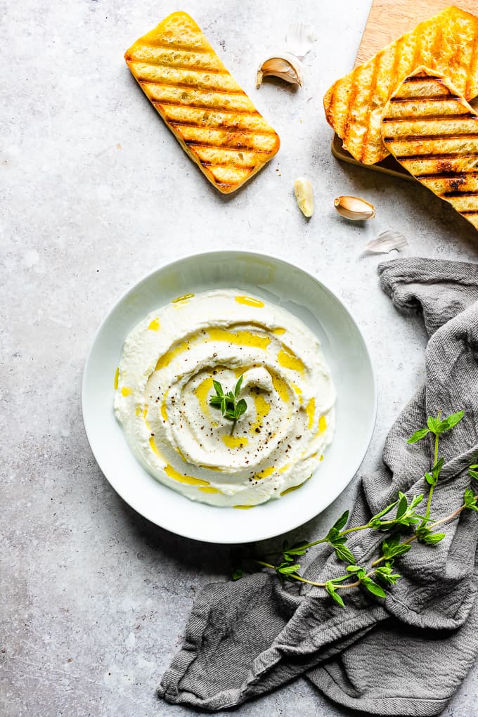Whipped ricotta in a white bowl topped with a drizzle of olive oil and a spring of oregano with grilled bread in the background.