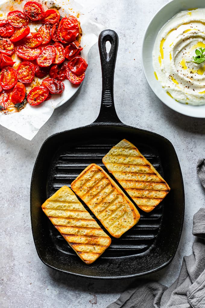 Three slices of bruschetta breads on a cast iron grill skillet with roasted tomatoes and ricotta in the background.
