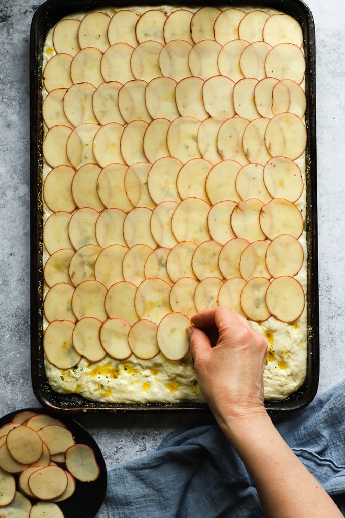 Topping focaccia bread with thin slices of potatoes in a sheet pan.