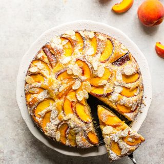Fresh peach and almond cake with one slice cut off, on a white cake stand.