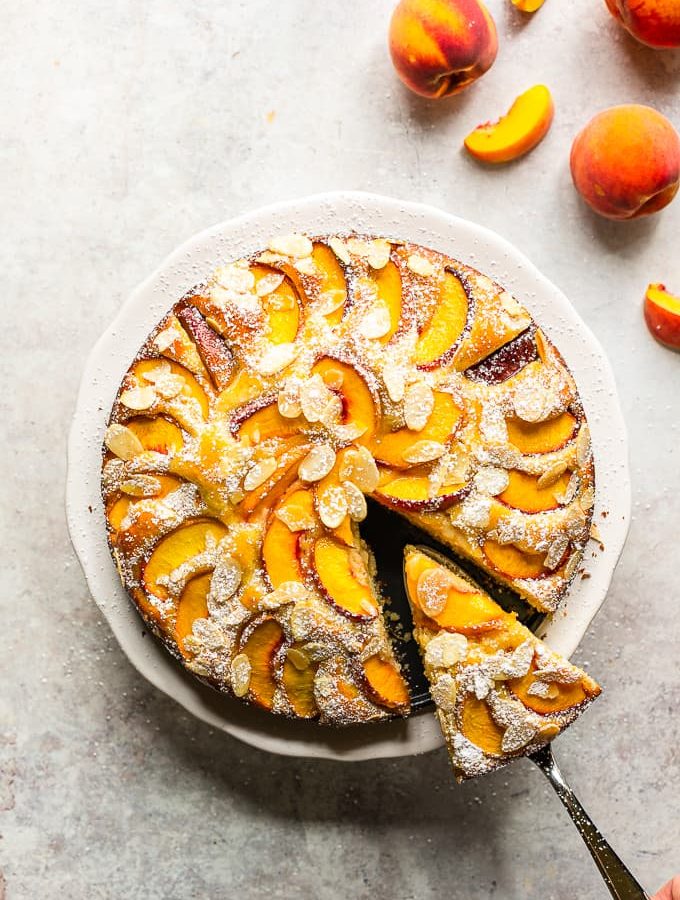 Fresh peach and almond cake with one slice cut off, on a white cake stand.